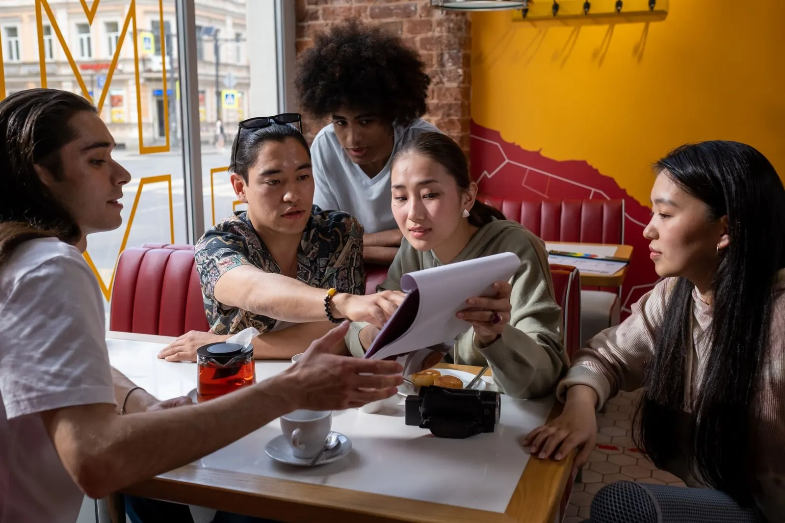 A group of friends talk around a cafe booth table, one holding a notepad.