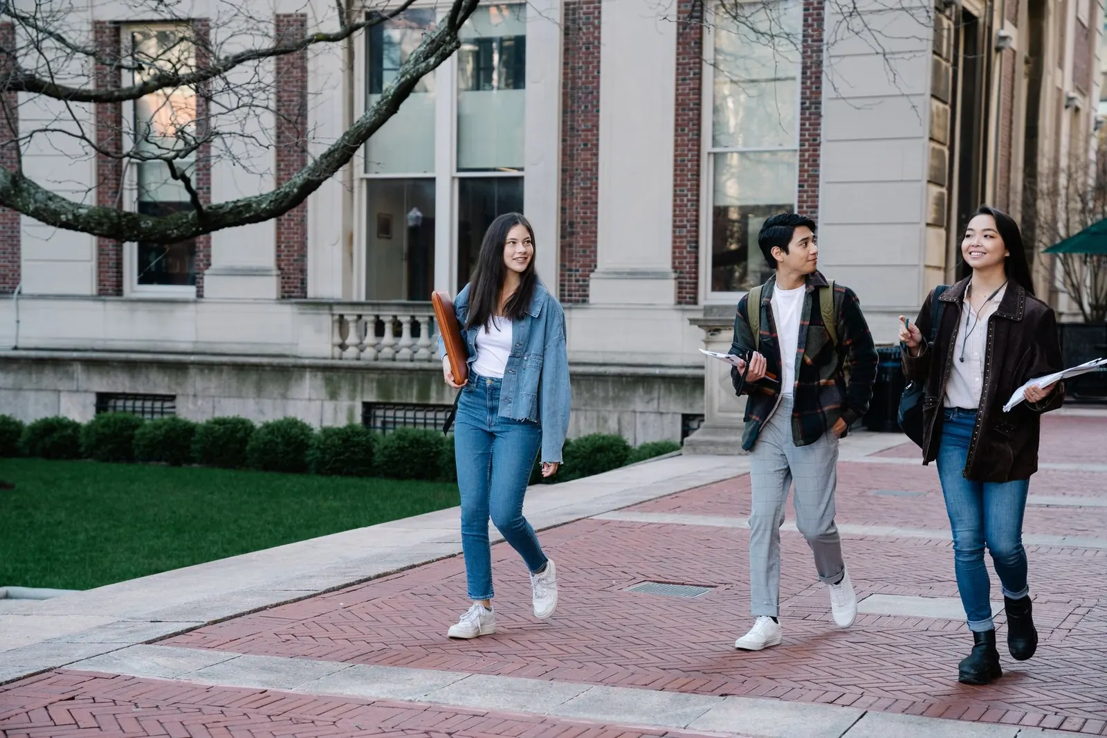 Three students walk along a brick path past a stone campus building and a green lawn.