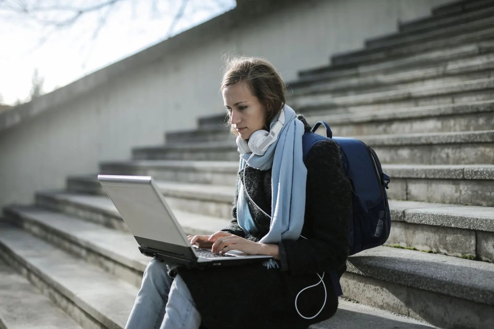 A student with headphones around her neck works on a laptop on outdoor concrete steps.