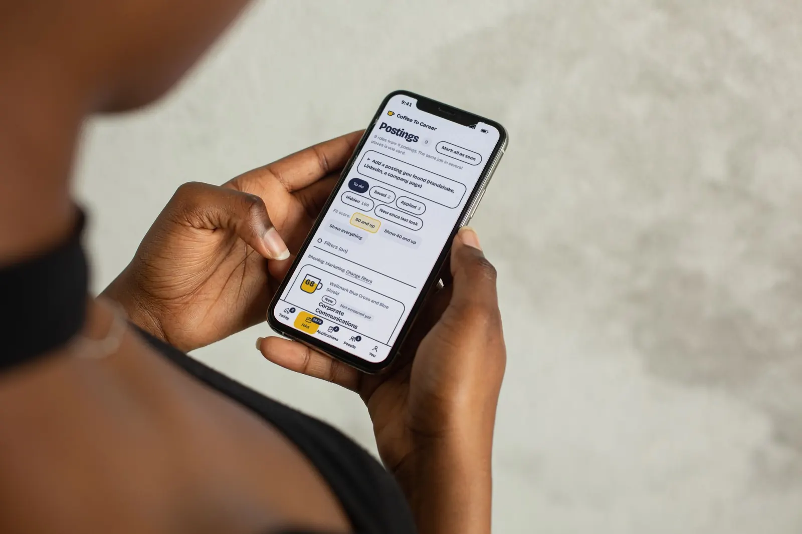 Close-up of hands holding a phone open to the Coffee To Career postings list, with sample data.