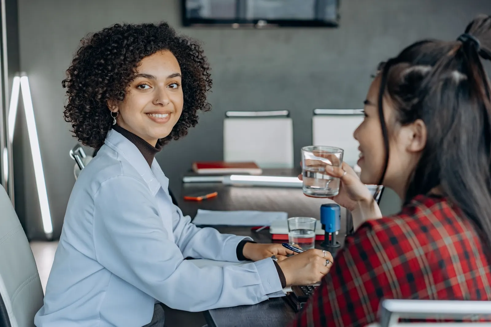 A smiling young woman talks with a colleague across an office table.