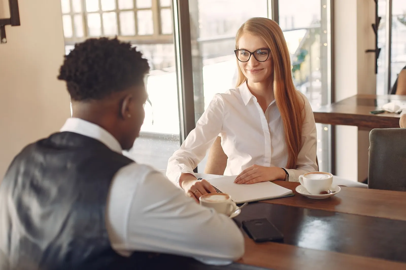 A young man and a woman in a white blouse talk over coffee at an office table by a window.