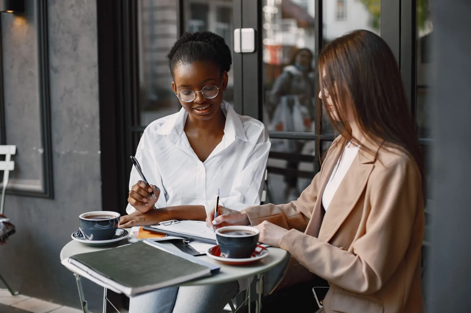 Two women write in notebooks at a small cafe table with coffee.