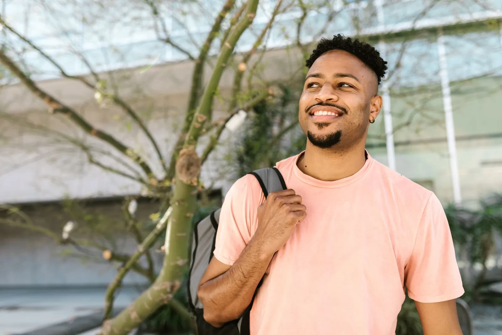 A smiling student in a peach T-shirt with a backpack strap over his shoulder, outside on a sunny campus.