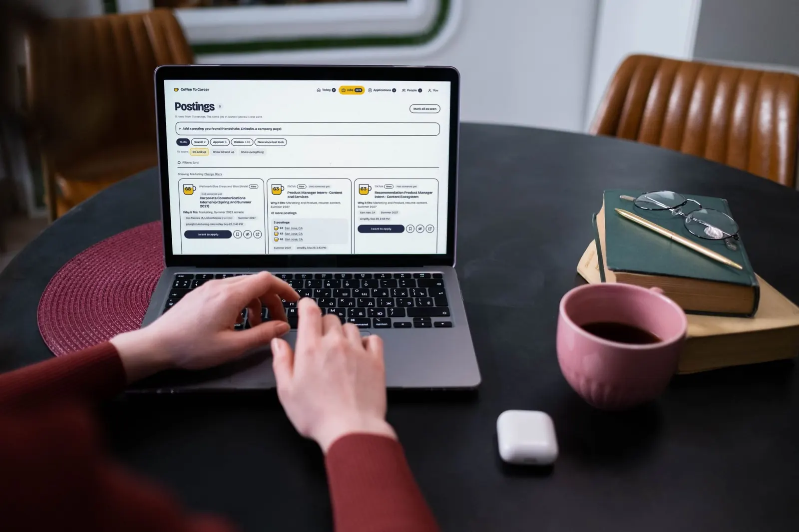 A student types on a laptop at a round black table, the Coffee To Career postings page on screen with sample data, a pink mug and books beside it.