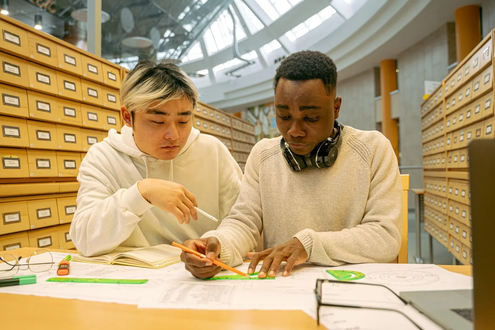 Two students in a library work through notes together at a table lined with card catalog drawers.