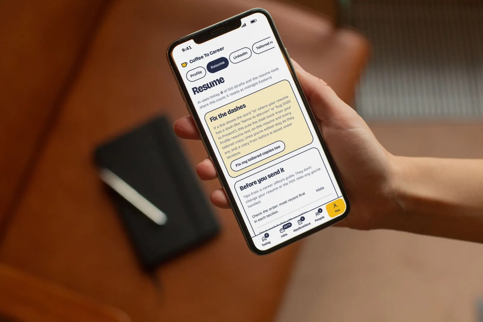 A hand holds a phone showing the Coffee To Career resume page with sample data, a notebook on a leather chair behind.