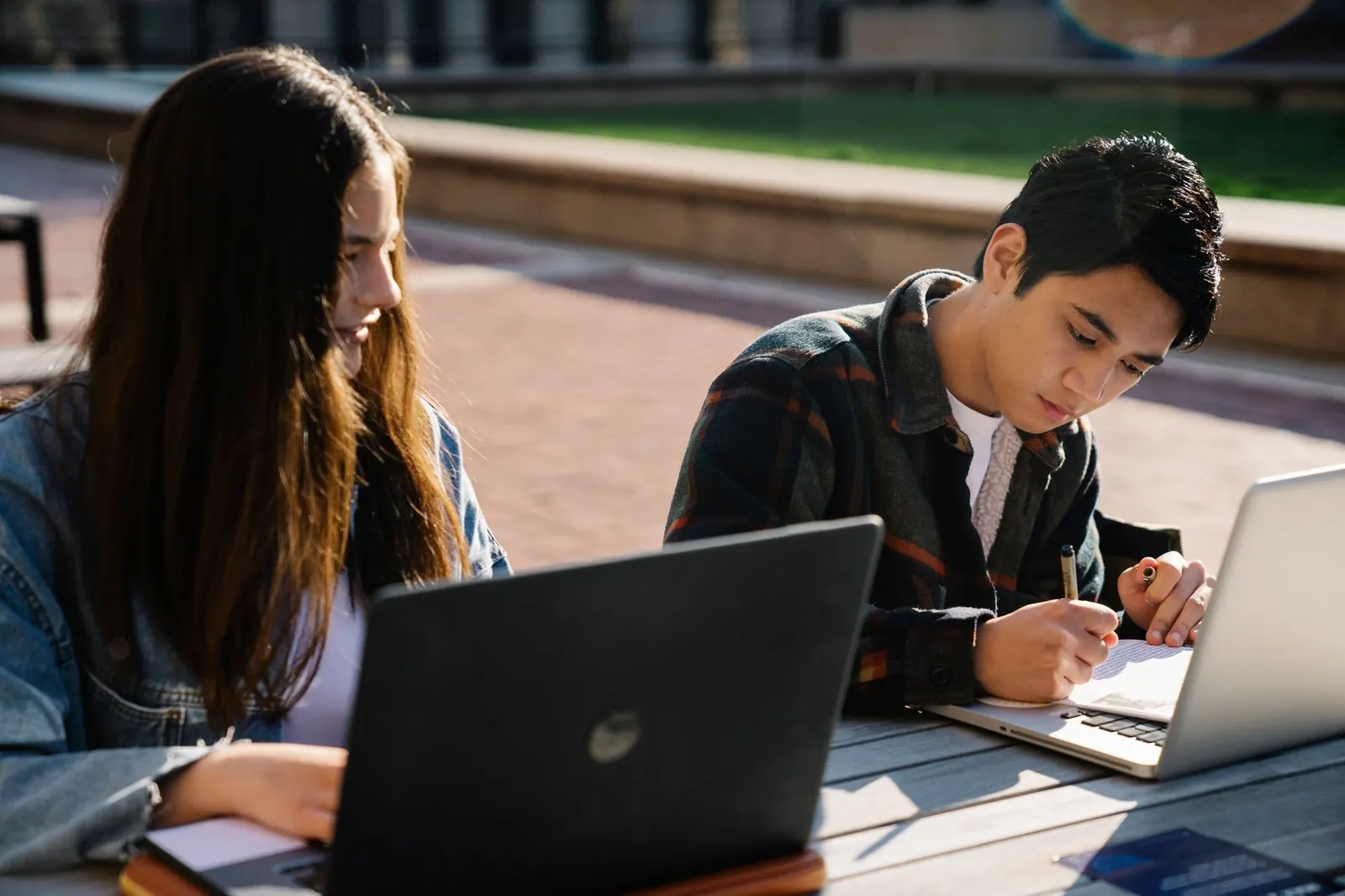 Two students work side by side at an outdoor table, one writing notes, laptops open.
