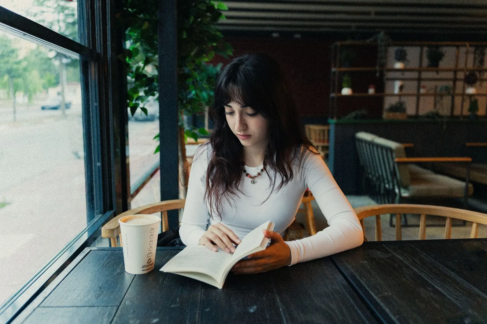 A young woman reads a book at a cafe table by a large window, a paper cup beside her.