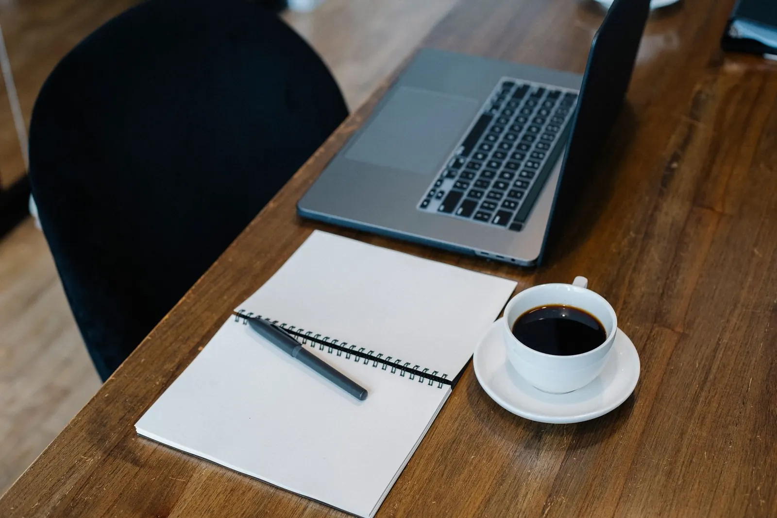 A laptop, an open notebook and a cup of black coffee on a wooden desk, seen from above.