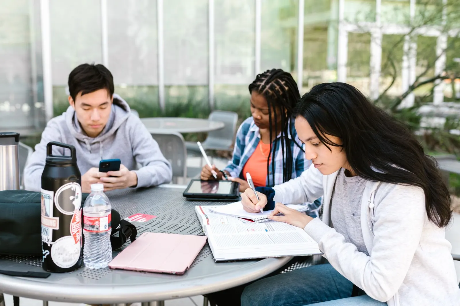 Three students study at an outdoor table with books, a notebook and water bottles.